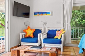 Una habitación con dos sillones blancos con cojines azules, un televisor de pantalla plana en la pared, un cuadro pequeño y una ventana con cortinas blancas.