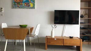 Een moderne woonkamer met een houten eettafel, stoelen, een flatscreen televisie aan de muur en een boekenkast met decoratieve objecten.