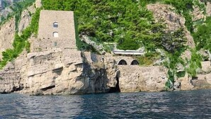 Un edificio costiero con una torre in pietra e finestre ad arco, immerso nel verde e con vista sul mare.