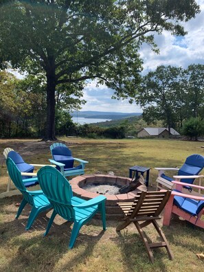 A fire pit surrounded by Adirondack chairs in a grassy outdoor area with a view of a lake and hills in the background.