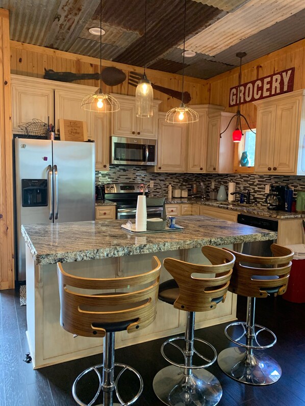 A kitchen with wooden cabinets, a stainless steel refrigerator, a microwave, and a granite countertop. There are three bar stools and a hanging light fixture.