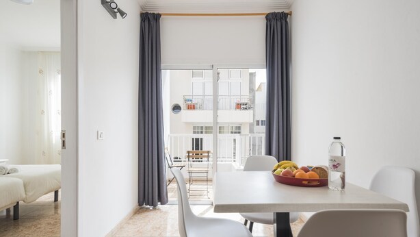 A modern apartment with a dining area, a balcony with a view of the building, and a bed in the background.