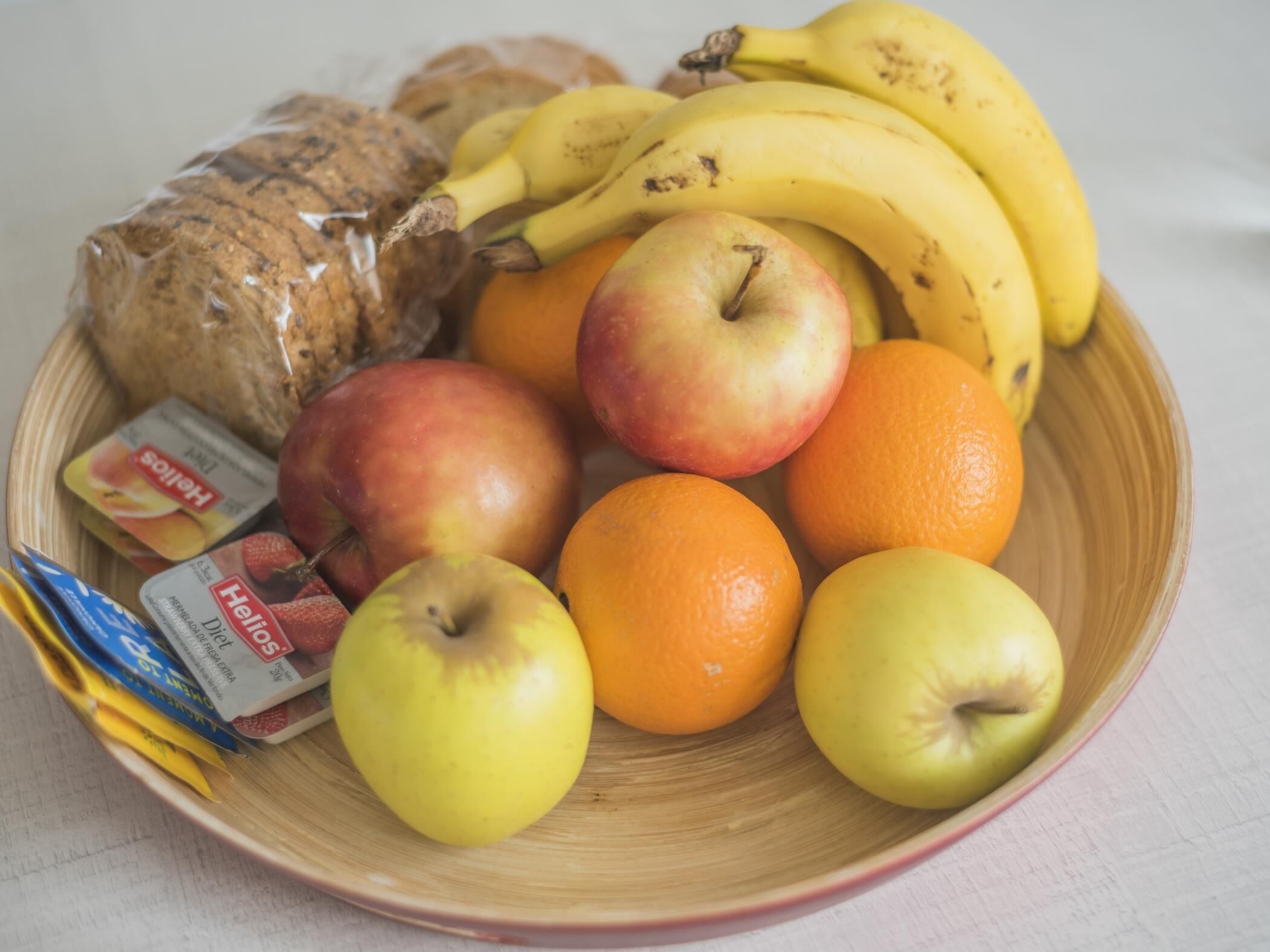 A bowl containing apples, oranges, bananas, a loaf of bread, and a package of Heloios yogurt.