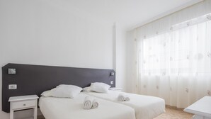 A hotel room with two beds, a nightstand, a desk, and a window with sheer curtains.