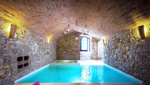 Une piscine intérieure aux murs de pierre, avec une eau d’un bleu limpide, un banc en bois agrémenté de coussins et un escalier menant à une porte.