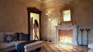 Un salon doté d’un sol à carreaux, d’un canapé blanc, d’une cheminée avec des bougies allumées et d’un miroir encadré d’un cadre orné.