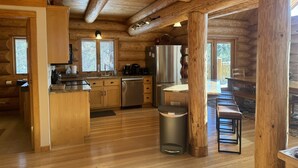 Interior de una cabaña de troncos con una zona de cocina que incluye armarios de madera, un refrigerador de acero inoxidable y una mesa de comedor con bancos.