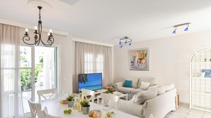 A bright living room with a white sofa, a dining table with chairs, a TV, and a chandelier.