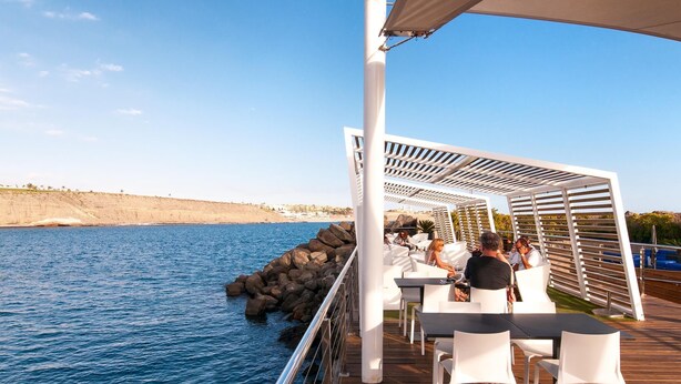 A deck with outdoor seating overlooking a body of water, featuring a pergola structure and a rocky shoreline in the background.