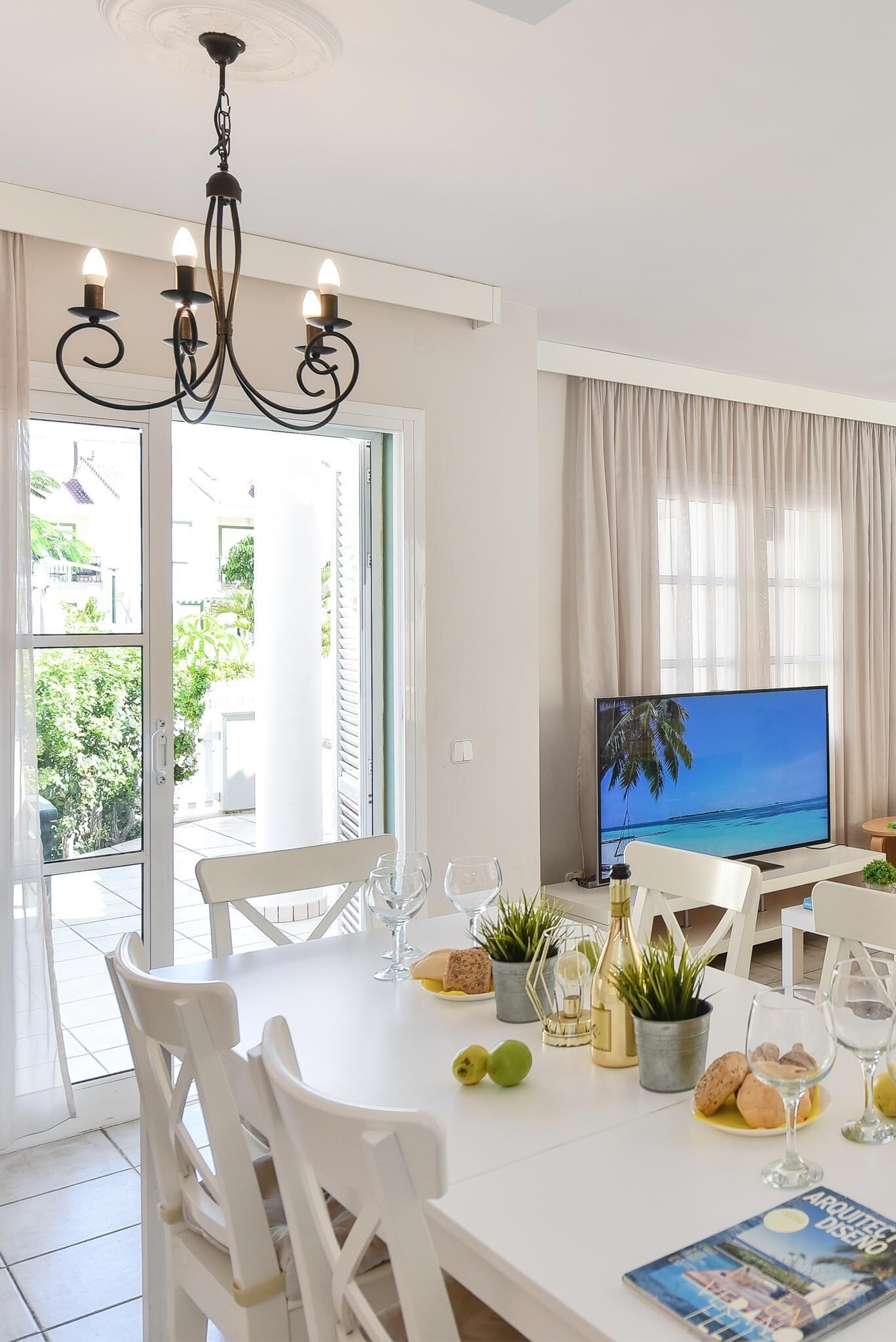 Un comedor con una mesa blanca, sillas, una araña y un televisor mostrando una escena de playa, además de vistas a la vegetación exterior.