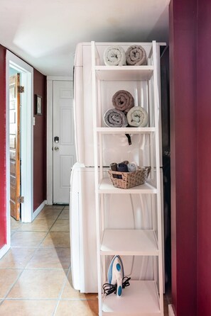 A white built-in shelving unit with rolled towels and a basket, a white refrigerator, and an ironing board with an iron.