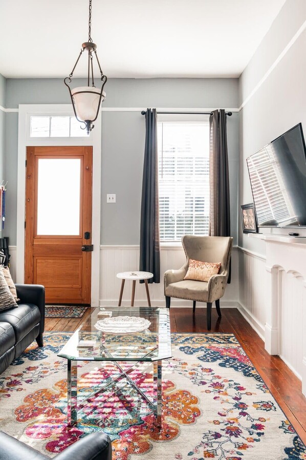 A living room with a glass coffee table, a leather sofa, a beige armchair, a patterned rug, a wooden door, and a window with curtains.