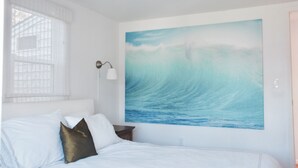 A bedroom with a large ocean wave painting on the wall, a bed with white and dark pillows, a nightstand, and a window with blinds. - Hamptons Waterfront Beach Home With Dock (Southampton)