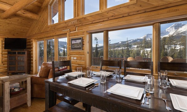 Una cabaña de madera con una mesa de comedor preparada para cuatro, amplias ventanas que ofrecen vistas a la montaña y un televisor instalado en la pared.