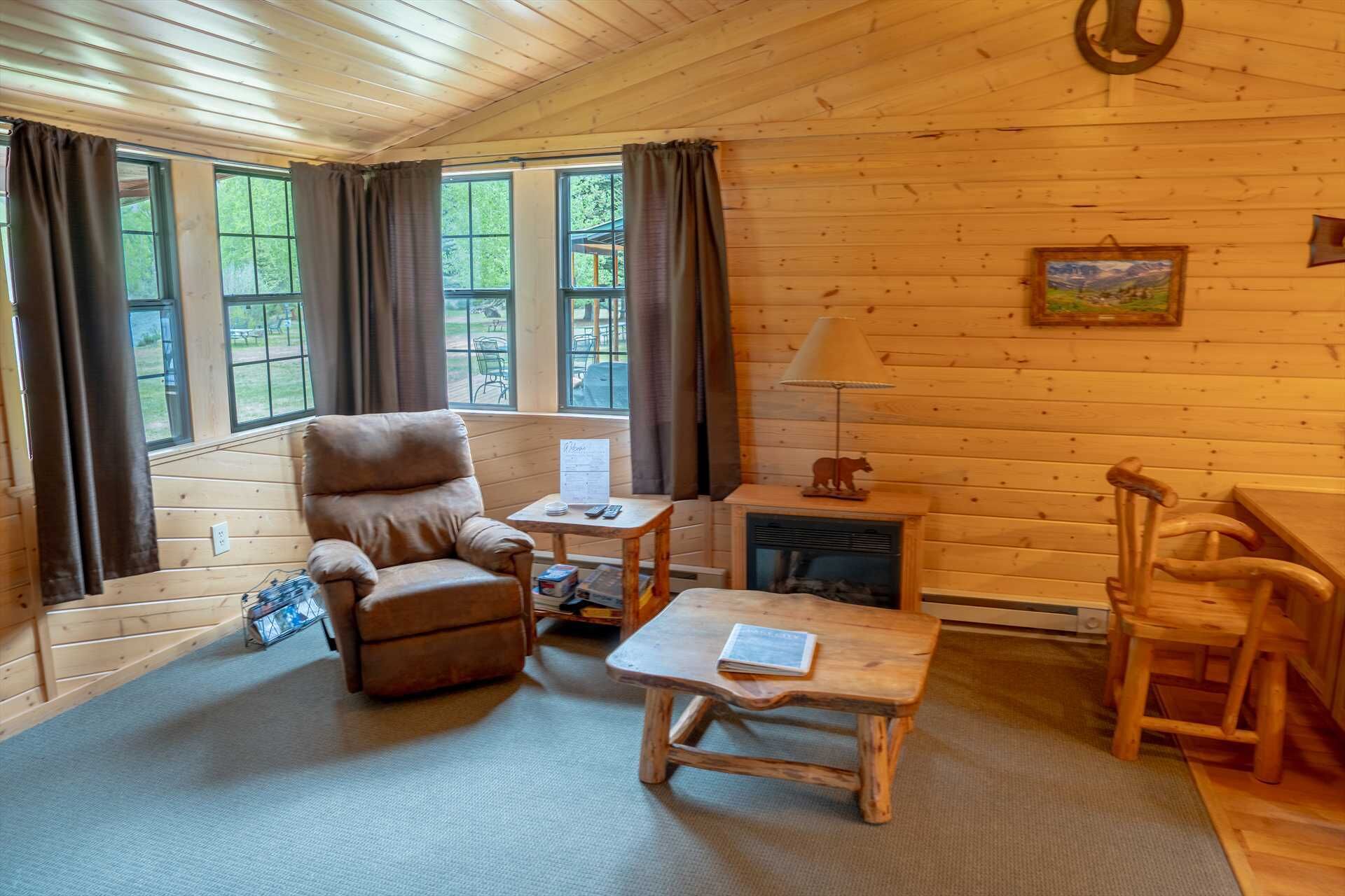 A cozy cabin interior with a wooden chair, a small table, a fireplace, and large windows with curtains.