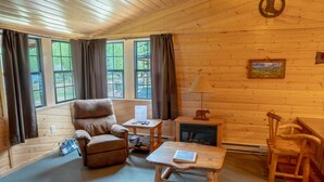 Interior acogedor de una cabaña con una silla de madera, una mesita, una chimenea y ventanas grandes con cortinas.