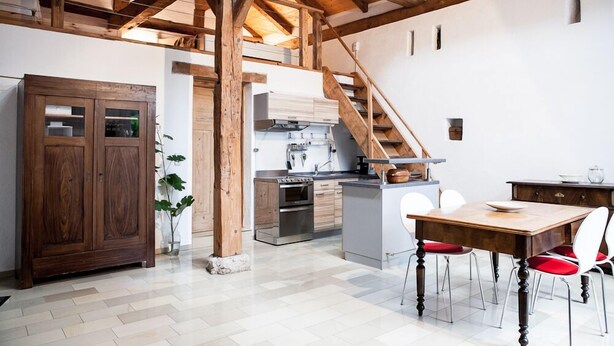 A spacious kitchen with wooden beams, a dining area with a wooden table and red chairs, and a staircase leading to an upper level. - Apartment/studio (converted barn, 40 sqm) with gallery (Schernfeld)