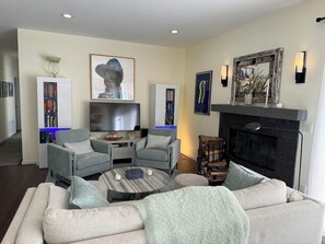 Un salon comprenant un canapé, deux fauteuils, une table basse, une cheminée, un téléviseur, une bibliothèque et des œuvres d’art décoratives accrochées au mur.