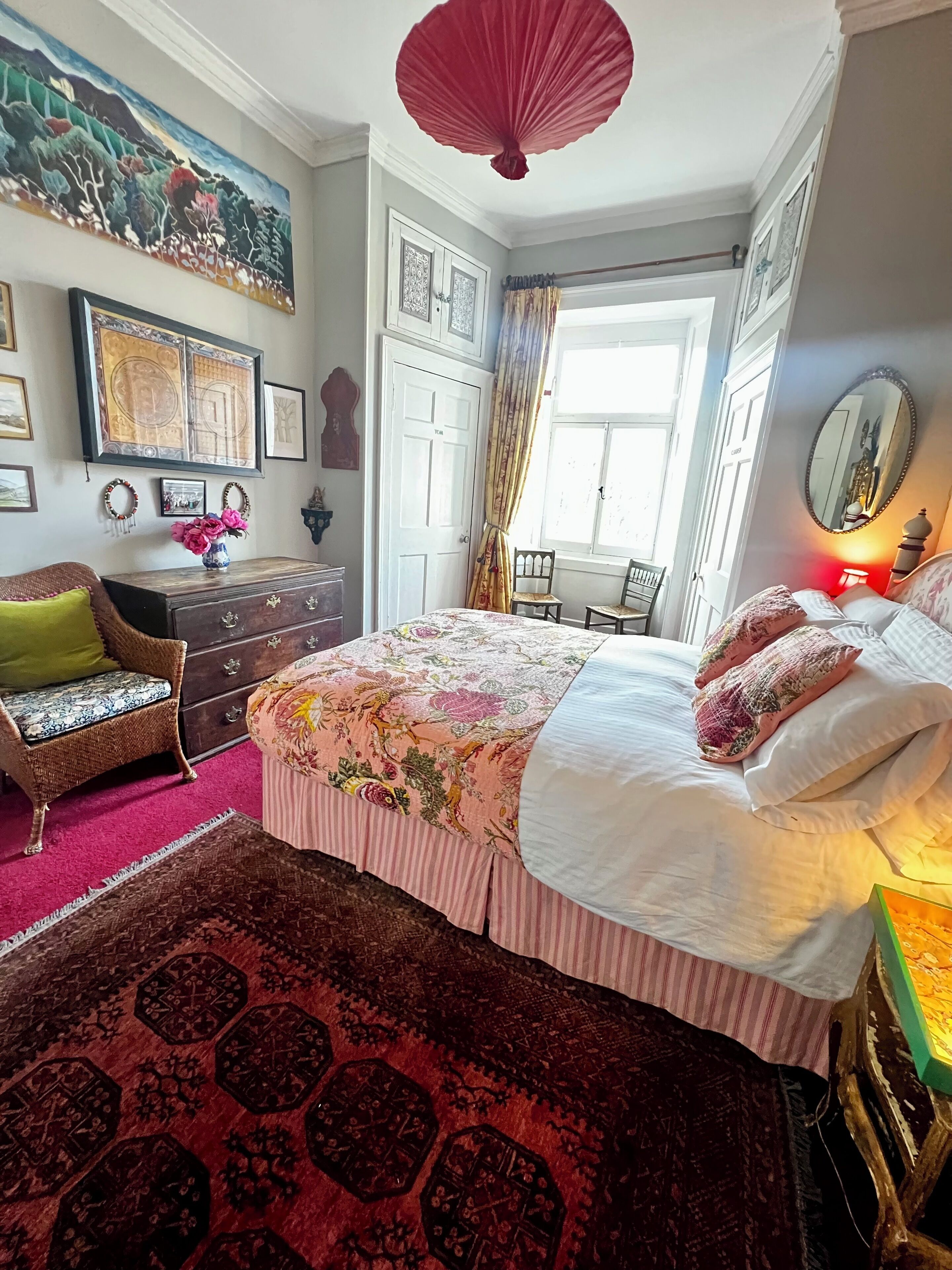 Een slaapkamer met een groot bed, een dressoir, een stoel, een raam met gordijnen, en een schilderij aan de muur.