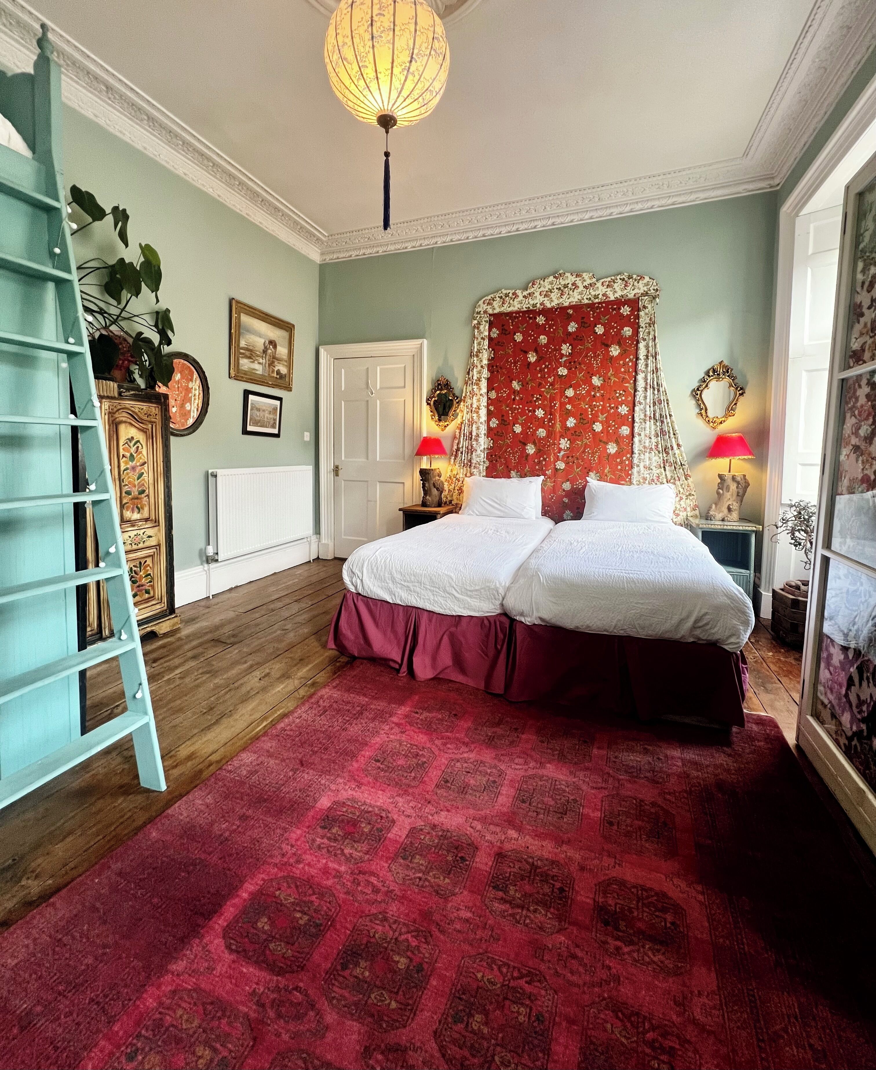 A bedroom with a double bed, a red patterned rug, a ladder, a white radiator, and a ceiling light.