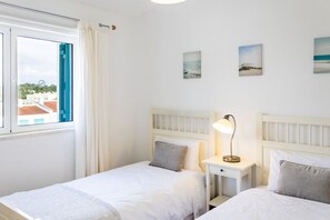 A bedroom with two beds, a window with curtains, and a nightstand with a lamp.