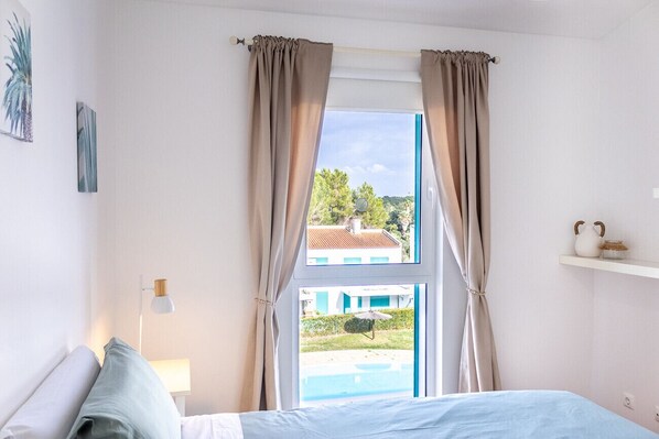 A bedroom with a bed, a nightstand, a lamp, a window with curtains, and a view of a pool and buildings outside.
