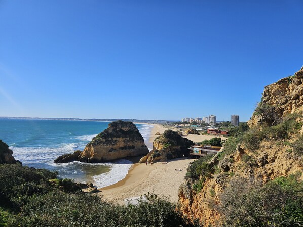 A coastal landscape with rocky cliffs, a sandy beach, and a clear blue sky.