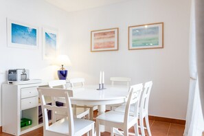 Un coin repas aux tons clairs, avec une table ronde, des chaises blanches, un vase bleu et des œuvres d’art encadrées accrochées au mur.