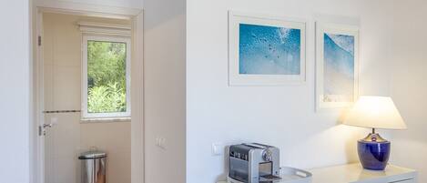 A small, clean room with a window, a trash bin, a coffee machine, a desk, a chair, and two framed pictures on the wall.