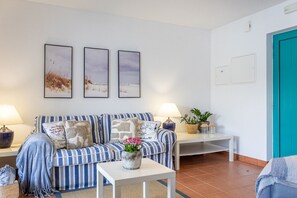 Un salon avec un canapé à rayures, deux tableaux accrochés au mur, une petite table sur laquelle est posé un vase de fleurs et une porte de couleur turquoise.