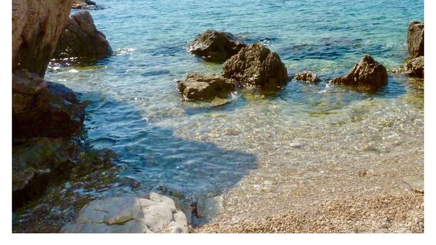 Paesaggio costiero con acque limpide, formazioni rocciose e un'isola in lontananza.