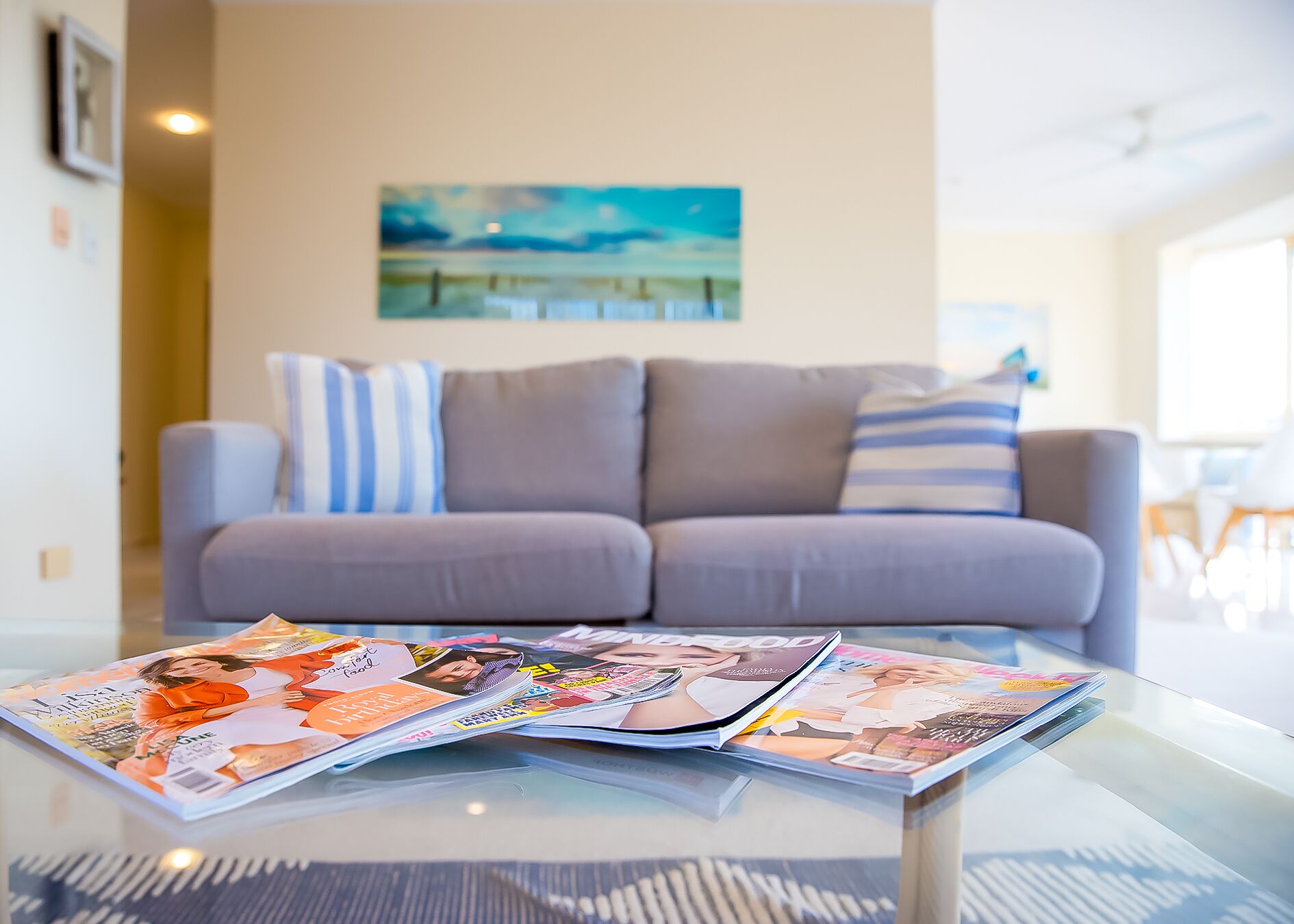 Una sala de estar con una mesa de centro de vidrio que muestra revistas, un sofá gris con almohadones a rayas y un cuadro en la pared.
