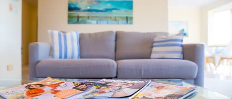 Una sala de estar con una mesa de centro de vidrio que muestra revistas, un sofá gris con almohadones a rayas y un cuadro en la pared.