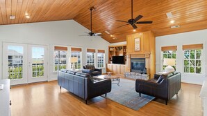 A spacious living room with a large sofa, a smaller sofa, a coffee table, and a fireplace. There are multiple windows with blinds, a ceiling fan, and a kitchen area visible in the background.