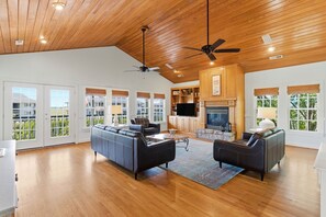 A spacious living room with a large sofa, a smaller sofa, a coffee table, and a fireplace. There are multiple windows with blinds, a ceiling fan, and a kitchen area visible in the background.