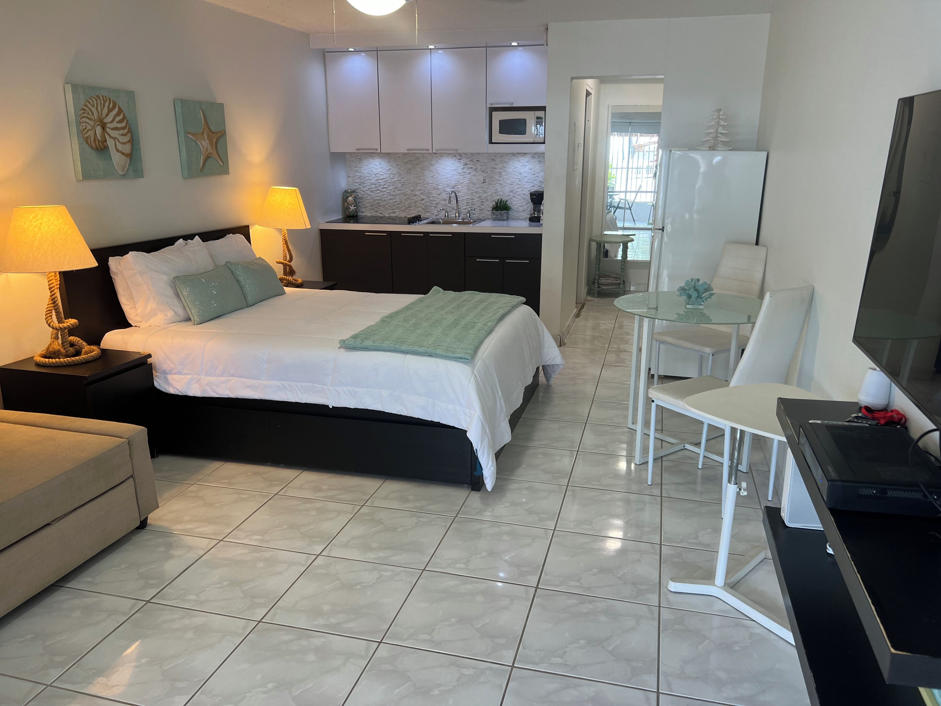 A hotel room with a bed, bedside tables, a desk, a chair, a TV, a kitchen area, and a dining area.