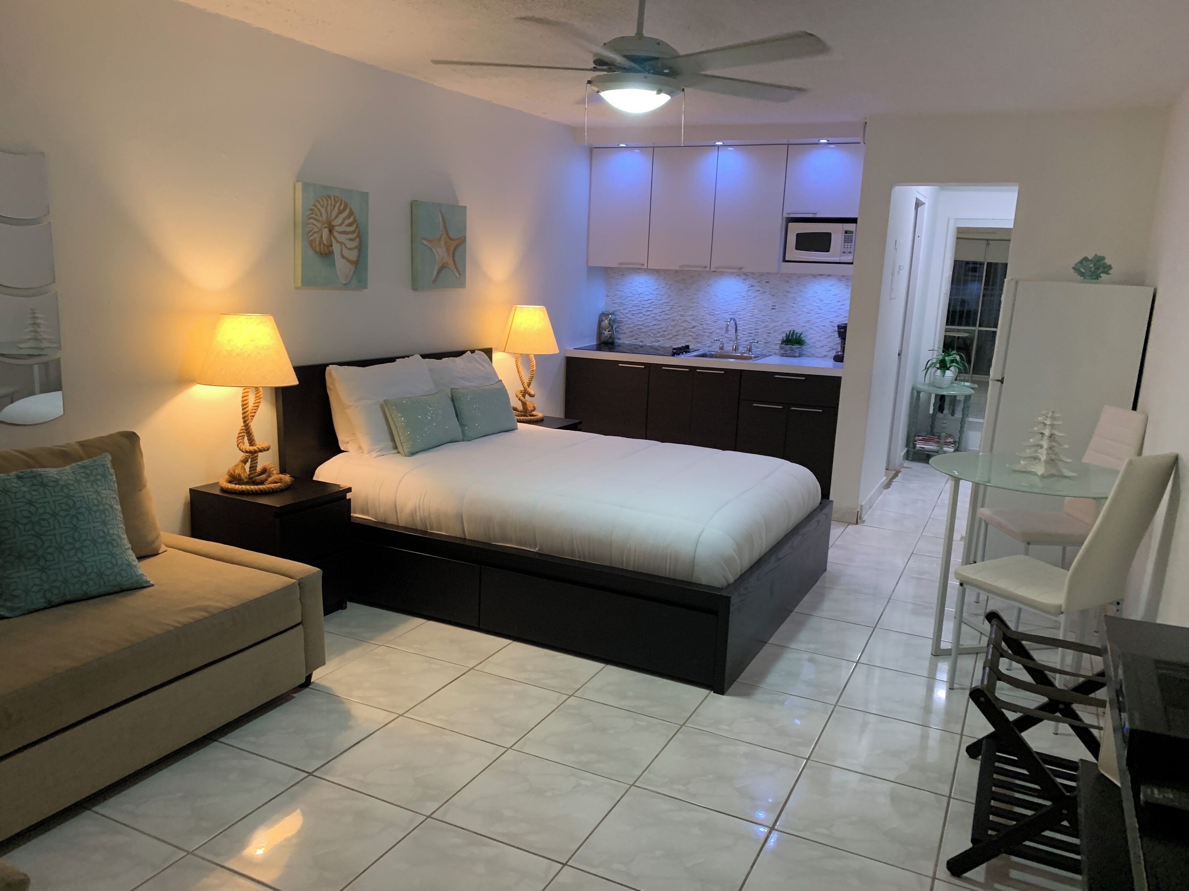 A bedroom with a bed, bedside tables, a nightstand, a ceiling fan, and a view into the kitchen area.