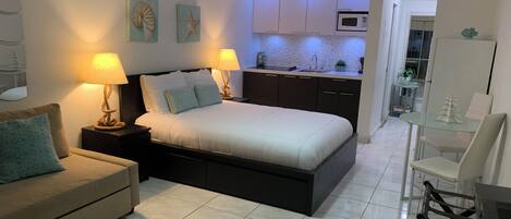A bedroom with a bed, bedside tables, a nightstand, a ceiling fan, and a view into the kitchen area.