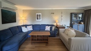 A living room with a blue sofa, a beige sofa, and a wooden coffee table. There is a brick wall with a framed picture and a cabinet with glass doors. - Parkview 1 Coastal 3 bedroom home,100m stroll to the beach, parks, cafés (Black Head)
