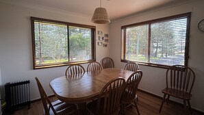 A dining room with a wooden table and chairs, large windows with blinds, a radiator, and a clock on the wall. - Parkview 1 Coastal 3 bedroom home,100m stroll to the beach, parks, cafés (Black Head)