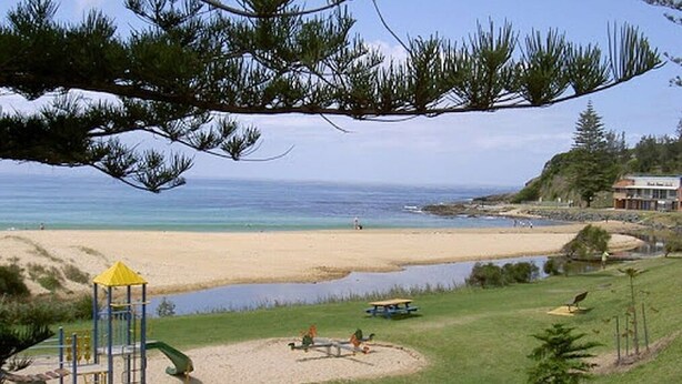 A beach with a playground, a small body of water, and a building in the background. - Parkview 1 Coastal 3 bedroom home,100m stroll to the beach, parks, cafés (Black Head)