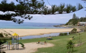 Une plage avec une aire de jeux, un petit plan d’eau et un bâtiment en arrière-plan.