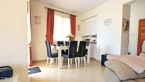 Uma sala de jantar com mesa e cadeiras, uma cozinha ao fundo e um relógio na parede.