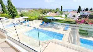 Piscina no terraço com água azul cristalina, rodeada por espreguiçadeiras e guarda-sóis, com vista para uma área residencial com casas e o mar.