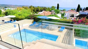 A rooftop pool with clear blue water, surrounded by lounge chairs and umbrellas, overlooking a coastal landscape with buildings and trees.