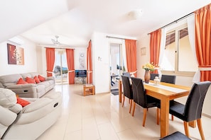 A modern living room with a sofa, dining table, and chairs. There is a TV and a small table with a vase. - Villa With Private Pool Hosting Up To 4 Guests - Great Sea Views. (Coral Bay)