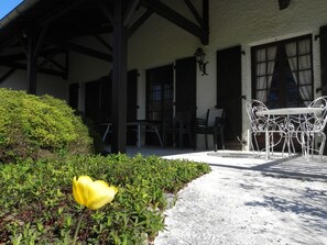 Une terrasse couverte avec une table et des chaises en métal blanc, une tulipe jaune au premier plan et un bâtiment avec des poutres et des fenêtres en bois foncé.