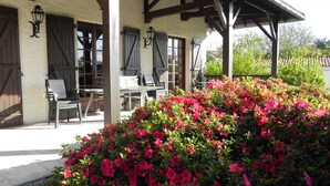 A covered outdoor seating area with tables and chairs, a white building with wooden beams, and a large bush of pink flowers. - GITE NOLAN *** SPA/SAUNA, 20MN FROM PUY DU FOU, IDEAL WITH FAMILY OR FRIENDS (Saint-Mesmin)