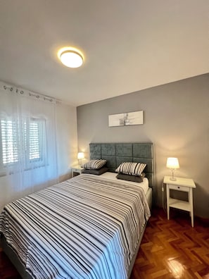 Un dormitorio con cama, dos mesitas de noche, una lámpara de noche, ventana con persianas y luz de techo.