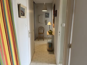 Um corredor estreito com piso de azulejos, uma cadeira de madeira, um abajur sobre uma mesinha e um objeto colorido listrado à esquerda.
